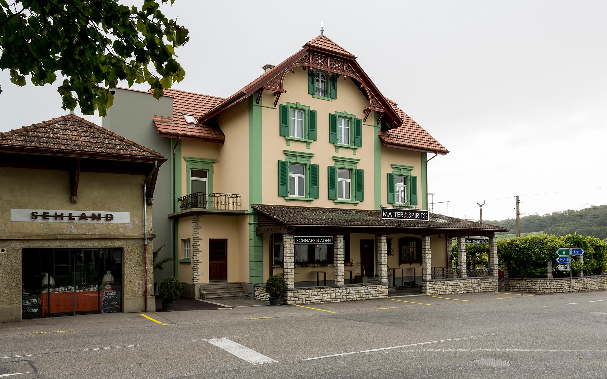 Aussenansicht des Gebäudes von Matter Spirits. Das renovierte Gebäude hat eine hellgelbe Fassade mit grünen Farbakzenten an den Seitenecken, passend zu den grünen Fensterläden.