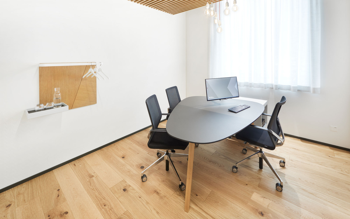 Dunkler Tisch mit hellen Beinen umgeben von 3 schwarzen Stühlen auf Rollen in lichterfülltem Raum