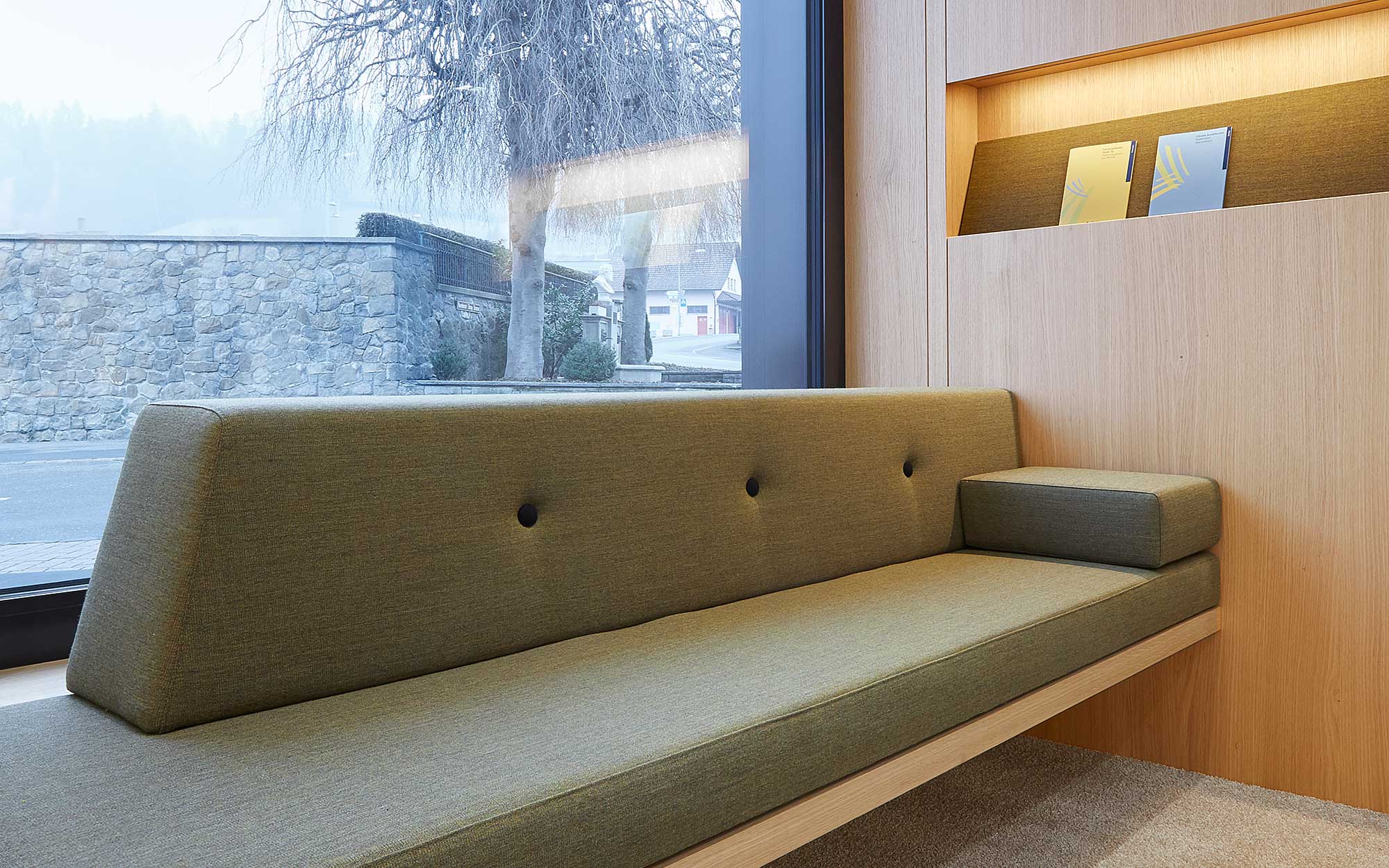 Die Wartezone vor dem Schalterbereich befindet sich vor einer grossen Fensterfront. Das Sofa ist aus einem naturnahen, abgedunkelten grünen Stoff gefertigt.