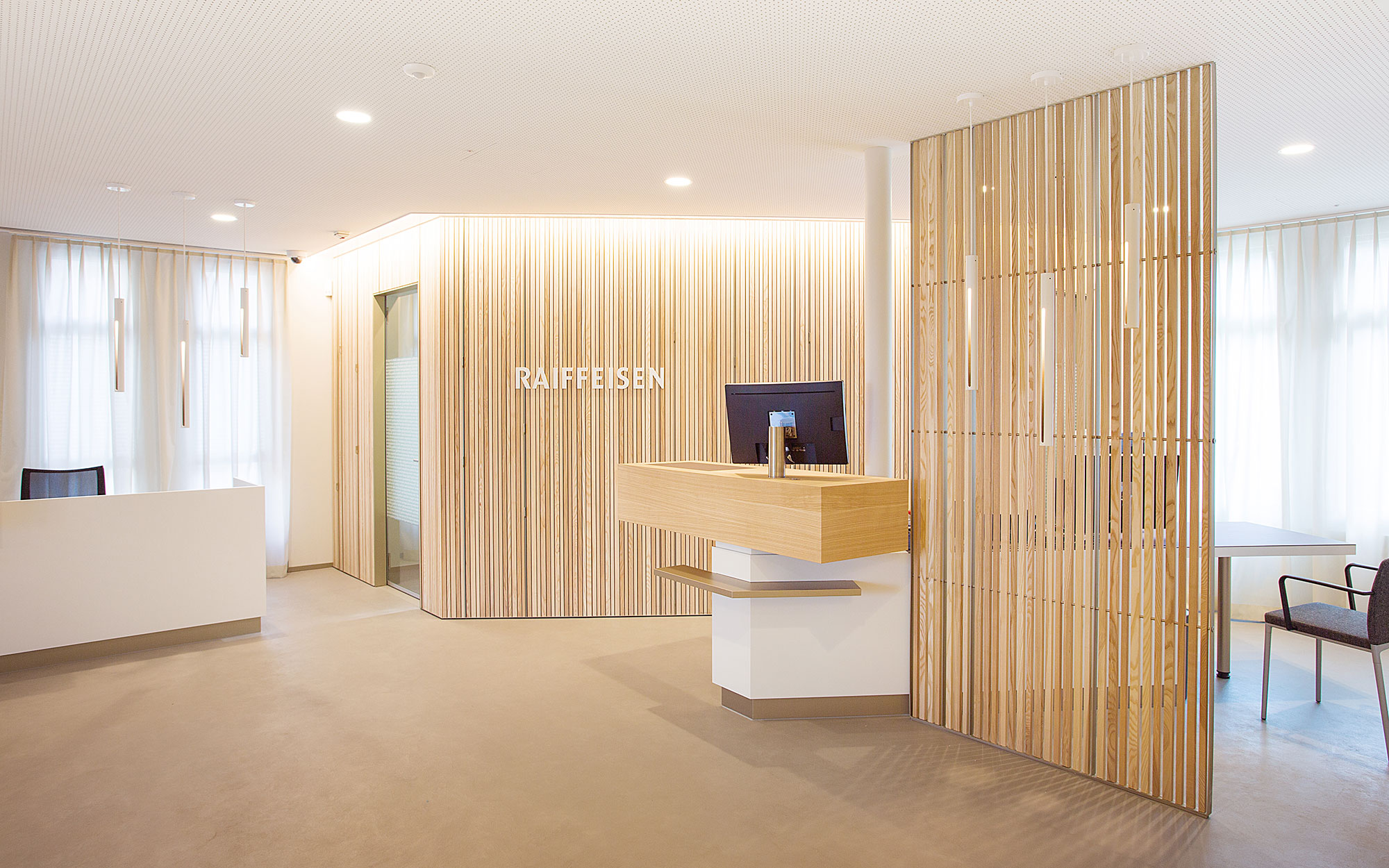 Beraterdesk in der Raiffeisenbank. Eine Trennwand aus Holzlamellen schirmt ab und sorgt für Diskretion. Der Bereich ist lichtdurchflutet und hell.