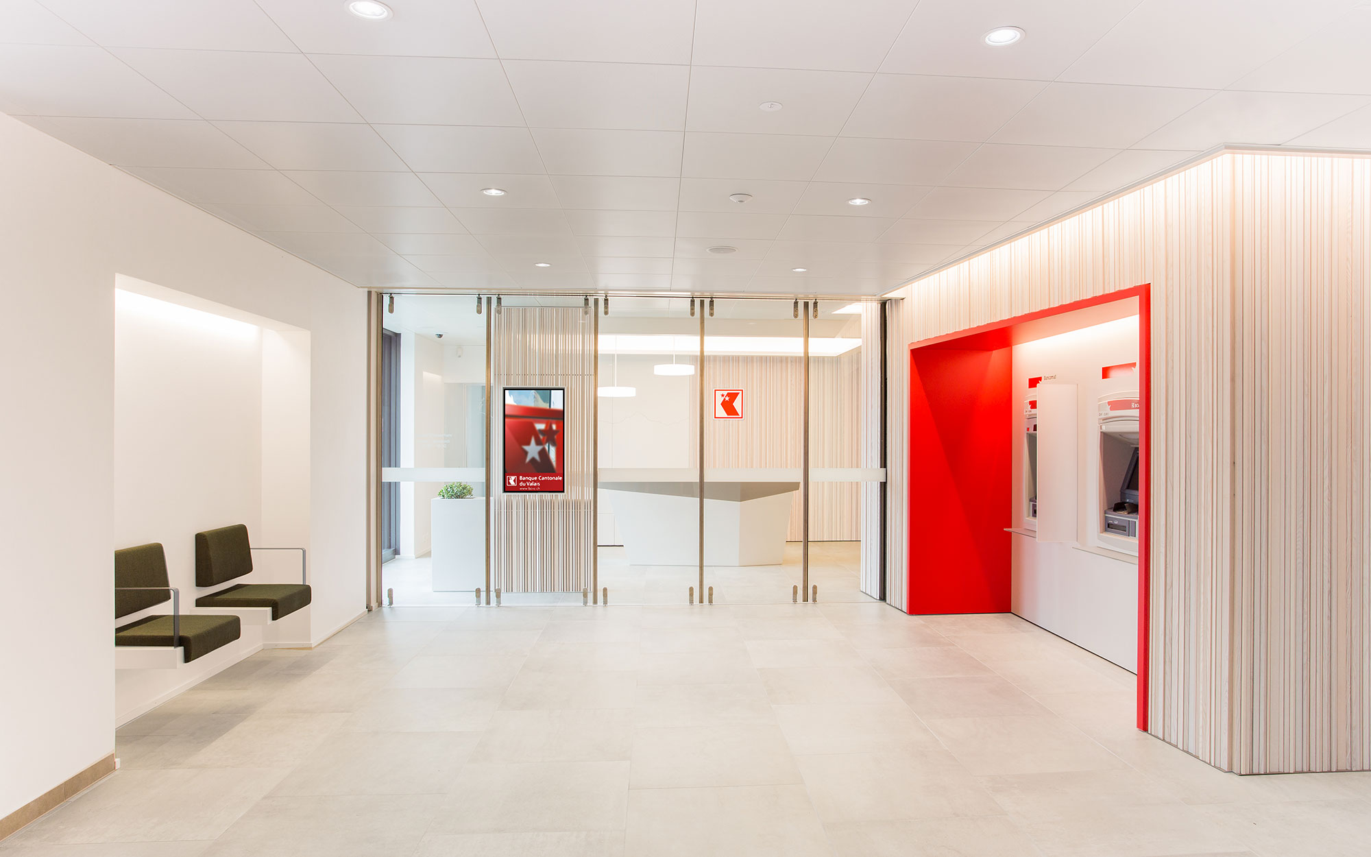 Eingangsbereich der Bank mit zwei Wartebänken vor der Bancomatenfront. Roter Rahmen fasst Bancomatenfront ein und ist in Holzlamellen-Wand integriert.